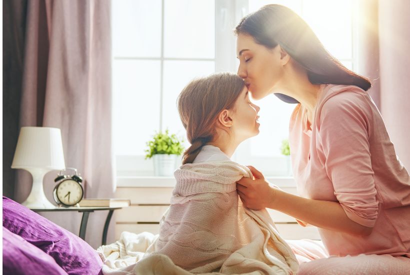 A mom in a warm sunlit room, kissing her daughter on the forehead when waking her up to symbolise a stress free, calm morning 